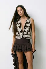 Chaleco de terciopelo marrón oscuro con escote en V pronunciado y bordados florales en blanco. Los bordes están decorados con un patrón geométrico en blanco.