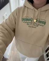 Canguro beige con capucha y bolsillo frontal tipo canguro. Presenta un estampado gráfico en el pecho con dos puños cerrados y la inscripción 'TA' LINDO' y 'TA' FRESCO', junto con el logo de la marca.