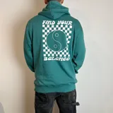 Canguro con capucha color verde, de corte regular, con estampado gráfico en la espalda que incluye un diseño de tablero de ajedrez, un símbolo de yin y yang y la frase 'Find your balance'.