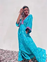 Túnica larga de estilo boho, color turquesa con estampado paisley en tonos azules y dorados. Presenta escote en V profundo con bordados decorativos en color fucsia y blanco, mangas amplias tipo kimono y ajuste en la cintura con cordón. El diseño incluye un ruedo con volado y terminaciones con borlas.