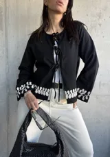 Chaqueta negra con escote redondo y cierre frontal con lazos. Presenta bordado de hojas blancas en los puños.