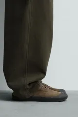 Zapatilla de cordones color verde militar, con parte superior de piel con acabado serraje y detalle de pespuntes en el empeine. Tiene horma redonda y suela plana de goma en color negro.