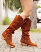 Bota estilo texana de caña alta, confeccionada en cuero gamuzado color marrón óxido. Presenta un diseño de corte en V en la parte superior de la caña, costuras decorativas y un taco bajo de madera.