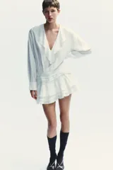 Vestido corto blanco de escote en V y manga larga con puños abotonados. Presenta volantes con bordados de encaje en el cuello, escote y falda. Cierre lateral con cremallera oculta.