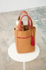 Matera de cuero color tostado con detalles bordados a mano en hilo rojo en las asas y en el bolsillo frontal. Cuenta con doble asa de mano y un llavero con borla colgante.