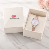 Reloj analógico de mujer con caja redonda de metal en tono oro rosa, esfera blanca con acabado brillante y malla metálica ajustable. Diseño minimalista y elegante.