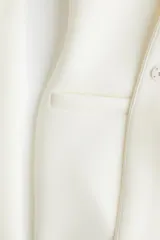 Blazer de mujer confeccionado en tejido efecto neopreno, en color blanco. Cuenta con cuello en V profundo, sin solapas, cierre frontal con broche a presión oculto, hombros caídos, mangas largas y bolsillos laterales de ojal simulados.