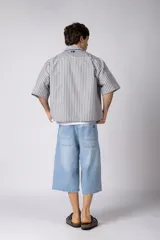 Camisa de manga corta de corte boxy, confeccionada en popelina a rayas celestes y blancas. Presenta cuello camisero clásico, cierre frontal con botones y estampado tipográfico en el pecho.