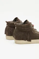 Zapatillas de caña media confeccionadas en cuero vacuno descarne color marrón. Presentan un diseño con flecos laterales, cordones al tono y un pequeño dije de corazón rojo en la parte superior de la lazada. Cuentan con suela de goma en color crema.