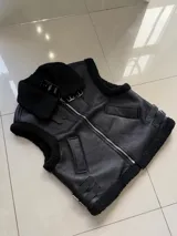 Chaleco de cuero sintético negro con interior y bordes forrados en corderito sintético. Presenta cierre frontal, cuello solapa, bolsillos laterales y correas ajustables con hebillas metálicas en los costados.