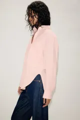 Camisa de corte oversize color rosa claro con diseño de rayas verticales sutiles. Presenta cuello solapa desmontable, cierre frontal con botones ocultos bajo solapa, mangas largas con puños y bajo asimétrico redondeado. Incluye dos bolsillos frontales amplios.