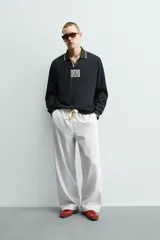 Polo de manga larga en tejido técnico acanalado color negro, con cuello solapa y abertura frontal. Presenta puños con ribete a contraste y una etiqueta aplicada en el pecho con el diseño conmemorativo de la Copa Mundial de la FIFA 1966.