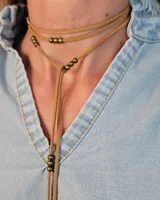 Collar estilo corbatín confeccionado en tiento de cuero color tostado, con diseño de doble vuelta y detalles de cuentas metálicas en tono bronce.