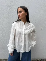 Camisa blanca de manga larga con textura a rayas verticales, canesú con volados y detalles de encaje en el pecho y el cuello alto. Cuenta con cierre frontal de botones ocultos y mangas abullonadas con volados en los puños.