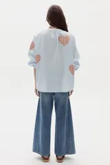 Camisa de algodón celeste a rayas verticales con parches estampados de corazones en tartán marrón y blanco. Posee cuello mao, cierre frontal parcial con botones y mangas largas abullonadas.
