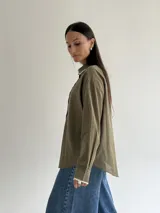 Camisa oversize de color marrón con cuello camisero y puños con detalle de bordado festoneado en color blanco. Presenta cierre frontal de botones y mangas largas.