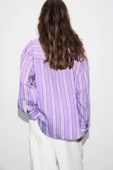 Camisa de corte oversize con diseño de rayas verticales en tonos violeta, blanco y bordó. Presenta cuello subido cruzado con cierre de botones frontal, mangas largas con puños y bajo asimétrico.