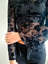 Remera de manga larga confeccionada en microtul transparente con diseño de flores aterciopeladas en color negro, cuello bote y costuras frontales visibles.