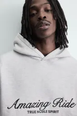 Sudadera con capucha y bolsillo frontal tipo canguro, confeccionada en tejido de algodón con interior perchado. Presenta diseño de corte boxy fit, mangas largas con puños y ruedo en rib, y texto bordado y estampado en el frente.