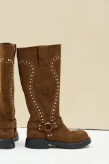 Bota alta de gamuza color marrón, con suela plana y gruesa, puntera redondeada y caña alta decorada con costuras en relieve y tachas plateadas. Cuenta con una tira con hebilla y argolla metálica en la base del empeine.