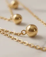 Set compuesto por un collar doble de cadena fina con dijes de esferas doradas y un par de aros tipo caravana con dije de esfera dorada, confeccionados en acero con baño de oro.