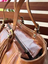 Cartera tipo tote de cuero color marrón tostado, con doble asa de hombro, detalles de hebillas metálicas doradas y flecos colgantes decorativos.