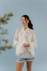 Blusa de algodón color blanco, con cuello alto y lazo frontal para atar. Presenta mangas largas con volados en los puños y un diseño holgado y romántico.