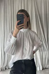 Camisa blanca de estilo romántico con textura de plumeti y rayas verticales. Presenta cuello alto con volados, cierre frontal con botones forrados y mangas largas abullonadas con puños ajustados.