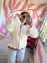 Campera de algodón premium color crudo con detalles en marrón y rosa. Presenta un cierre frontal, cuello redondo, mangas largas acampanadas y un logo bordado en el pecho.