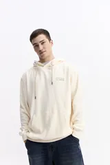 Canguro con capucha y cordones ajustables, de color crema con textura tipo waffle. Presenta un bolsillo frontal tipo canguro y un pequeño logo estampado en el pecho.