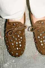 Zapato tipo náutico de serraje color marrón, con detalle de tachas metálicas plateadas en todo el cuerpo y cordones decorativos en la parte delantera. Presenta punta redonda y suela plana.