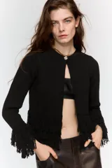 Chaqueta de punto color verde oscuro, con cuello redondo, manga larga y cierre frontal con un solo botón. Presenta detalles de flecos en el ruedo y en los puños.