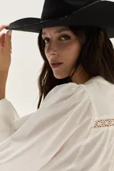 Blusa de algodón color blanco con mangas abullonadas, cuello mao y cierre frontal con botones. Presenta detalles de encaje tipo vainilla en el frente y en la parte superior del pecho.