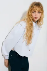 Camisa corta de sarga de algodón en color celeste, con cuello elevado, botonadura frontal con botones oscuros y corte trapecio. Presenta hombreras, mangas largas abullonadas con puños abotonados y detalles de frunces en los hombros.