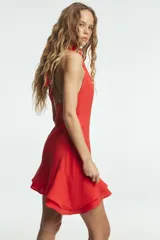 Vestido corto de tejido fluido en color rojo, con diseño de cuello halter y detalle de frunces laterales. Presenta un bajo asimétrico acabado en volantes y cierre lateral con cremallera oculta.