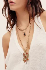 Collar largo de cadena dorada con un gran dije de corazón ornamentado con detalles grabados y cuentas rojas incrustadas, del cual cuelgan dijes pequeños con cuentas y colgantes metálicos.