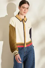 Cardigan de punto 100% algodón, diseño color block con cuerpo blanco, manga derecha verde militar y manga izquierda azul marino, ambas con tejido trenzado. Cuenta con cuello polo mostaza con ribete oscuro, cierre frontal con botones y detalles de rayas en el elástico inferior en tonos mostaza, marrón y fucsia.