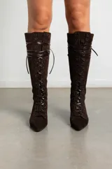 Bota alta de gamuza color marrón oscuro, con diseño de cordones frontales y cierre lateral. Presenta una silueta estilizada con punta fina y un taco bajo tipo kitten heel.
