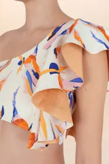 Top de bikini asimétrico, color blanco hueso, con estampado abstracto en tonos naranja, azul y rosado. Presenta un volado amplio en el hombro derecho y tirantes finos ajustables.