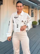 Camisa blanca de corte oversize y estilo cropped, con mangas largas y puños anchos. Presenta múltiples parches e insignias decorativas en el frente y las mangas, incluyendo detalles metálicos con cadenas.