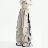 Pantalón deportivo color beige claro, estilo baggy o ancho, con cordón ajustable en la cintura y cierre completo impermeable (full zip) en los laterales. Presenta una franja vertical negra con tres líneas blancas a lo largo de la costura lateral, y un pequeño logo bordado en la parte superior del muslo.