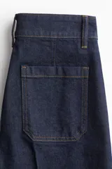 Pantalón de jean de denim oscuro, tiro alto, con pierna ancha y corte recto desde la cadera hasta el bajo. Presenta pliegues frontales y traseros, y un dobladillo ancho en los tobillos. Cierre frontal con dos botones dorados.