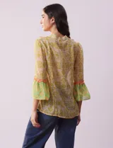 Blusa de gasa estampada con flores amarillas y verdes, marca Alex & Lili. Tiene cuello mao, mangas 3/4 acampanadas y cierre frontal con botones. Presenta volados verticales color naranja en el frente y en las mangas.