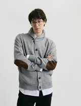 Cardigan de punto gris con cuello esmoquin, cierre frontal de botones y presilla superior. Presenta mangas largas con coderas en contraste color marrón y bolsillos de ribete.
