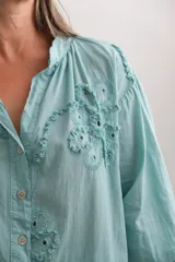 Blusa de color turquesa con cuello mao, cierre frontal de botones y mangas tres cuartos abullonadas. Presenta un diseño con bordados florales en relieve y detalles calados en el frente.