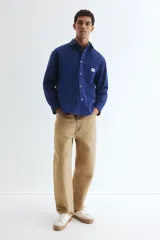 Sobrecamisa azul de lona gruesa de algodón, con cuello, tapeta con botones a presión y canesú en la espalda. Tiene mangas largas con botón a presión en los puños y dos bolsillos superiores de parche. El bajo es levemente redondeado y el corte es oversized.