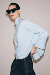 Camisa de corte oversize con estampado de rayas verticales, cuello redondo tipo mao y cierre frontal con botones. Presenta manga larga con puños anchos, un bolsillo de plastrón con solapa en el frente y bajo asimétrico.