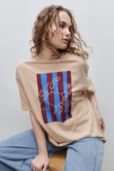 Remera blanca de calce relajado y silueta amplia, con cuello redondo, mangas cortas y estampa gráfica frontal de un cangrejo sobre franjas en tonos rosado y rojo.