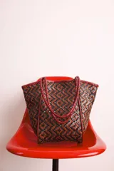 Bolso tipo tote confeccionado en cuero trenzado a mano, combinando tonos marrones y negros. Presenta asas trenzadas en color bordó y bordes reforzados en el mismo tono.