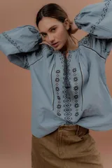 Blusa de estilo bohemio color celeste, con mangas abullonadas y amplias, escote redondo con tiras colgantes y borlas, y detalles de bordados geométricos en contraste de color en el pecho y las mangas.